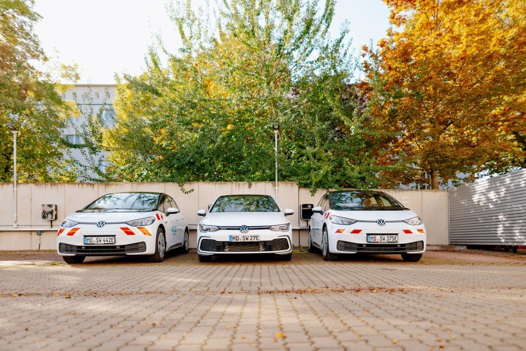 Drei weiße Elektroautos mit rot-weißen Warnstreifen auf den Stoßfängern, die nebeneinander auf einem Parkplatz stehen. Zwischen dem mittleren und rechten Fahrzeug befindet sich eine Ladesäule.